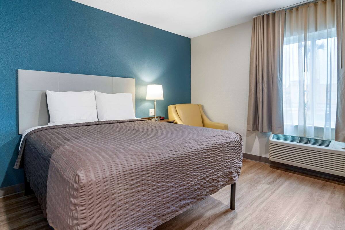 A hotel room with a bed, a chair, a lamp, and a window with curtains.