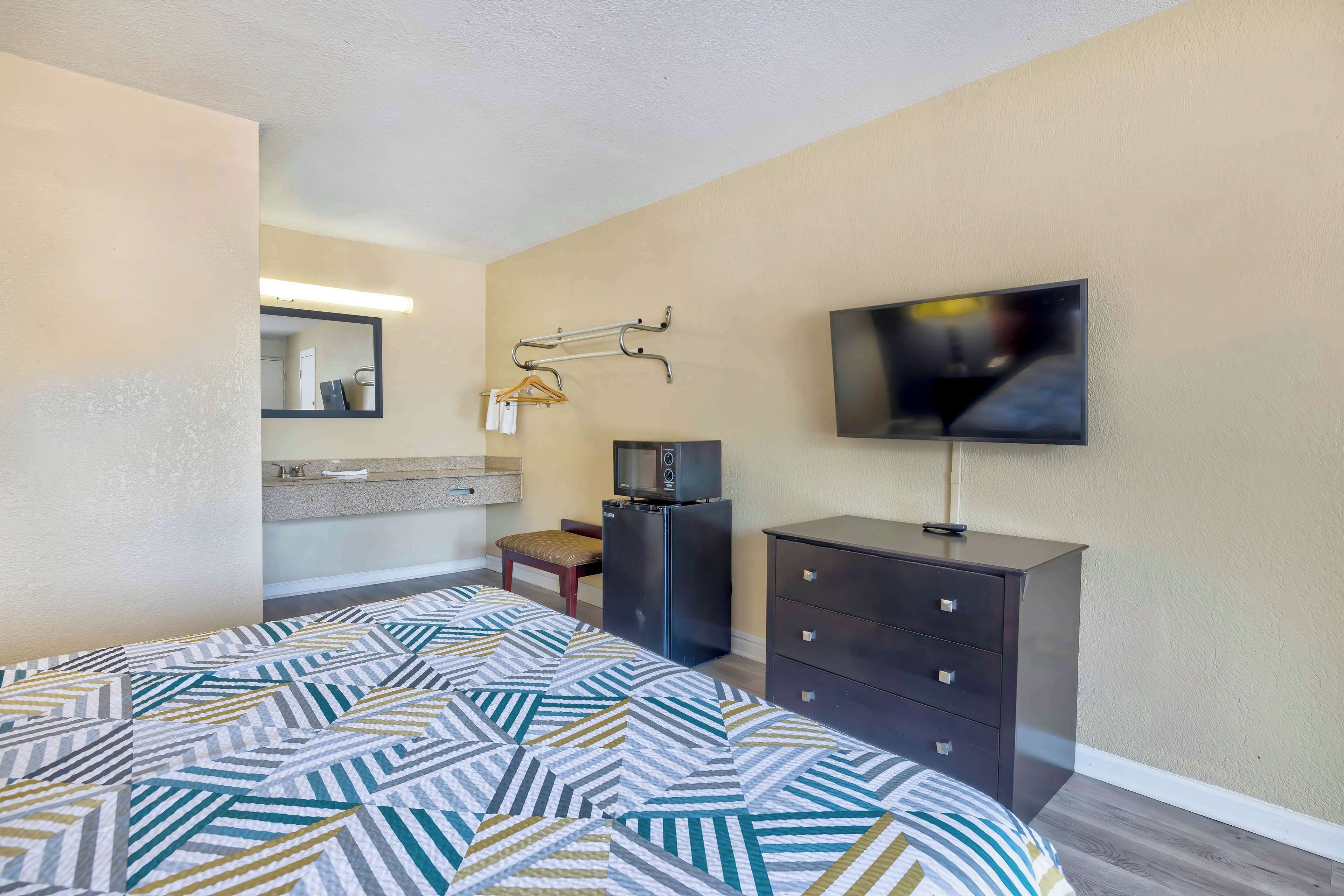A hotel room with a bed, a dresser, a television, a mirror, and a bathroom with a sink and towel rack.