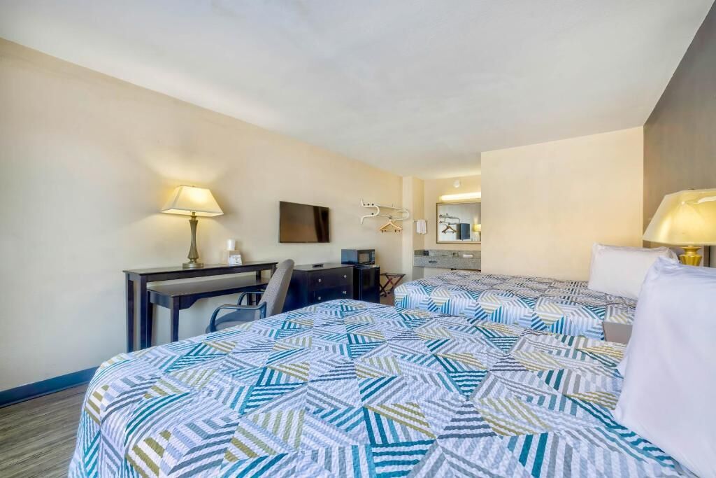 Une chambre d’hôtel avec deux lits, un bureau, une chaise, une lampe, un miroir et un porte-manteau fixé au mur.