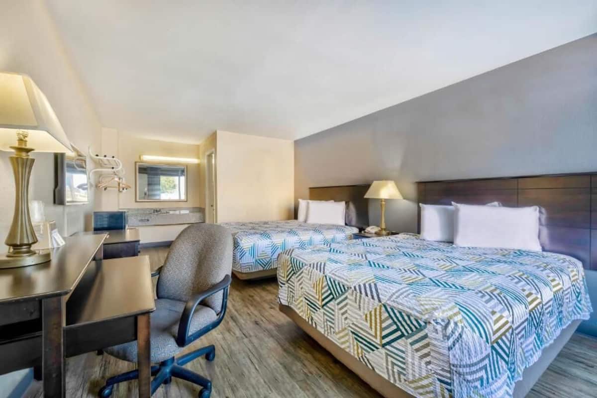 A hotel room with two beds, a desk, a chair, and a lamp.