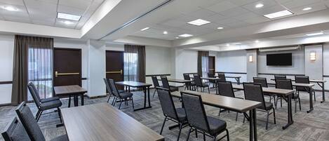 A conference room with multiple tables and chairs, a flat-screen TV mounted on the wall, and large windows with curtains.
