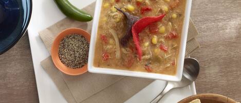 A bowl of soup with a red chili pepper on top, a small bowl of black pepper, and a wooden bowl of tortilla chips on a table.