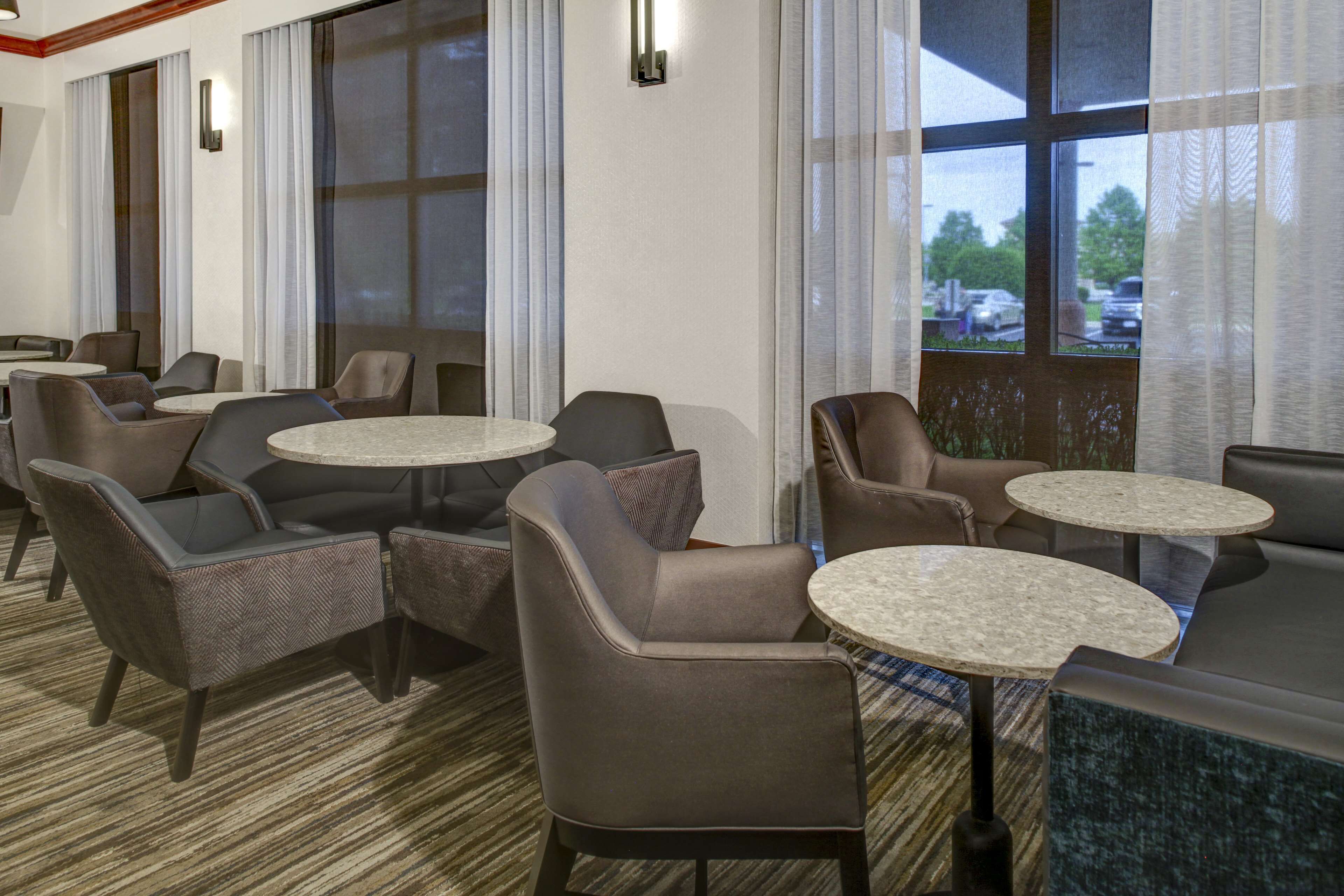 A hotel lobby with multiple seating areas, including sofas and round tables, and large windows with curtains.