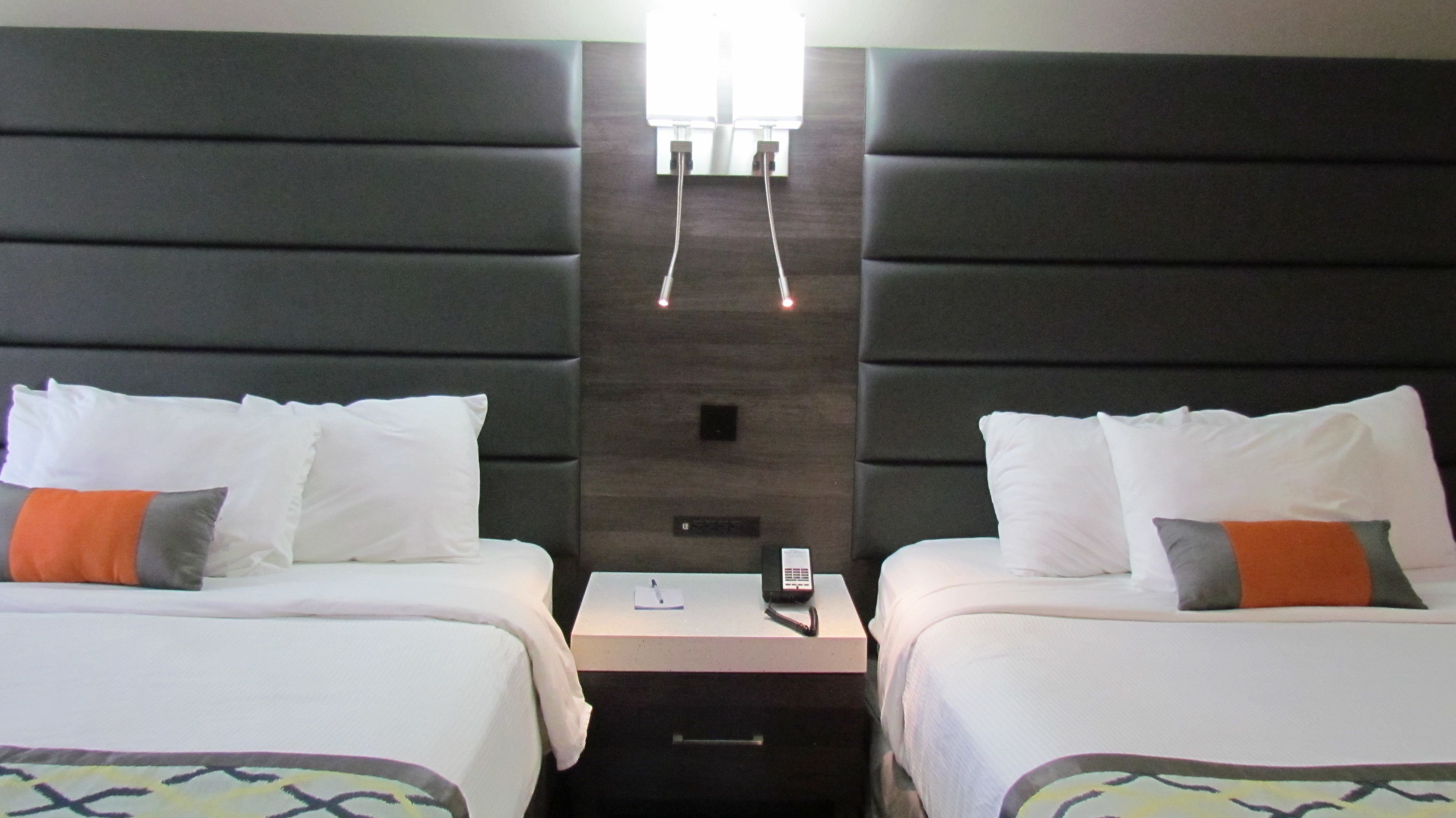 A hotel room with two beds, each with white bedding and orange pillows. There is a nightstand between the beds with a phone and a small item on it. The room has a dark headboard and a light fixture above the beds.