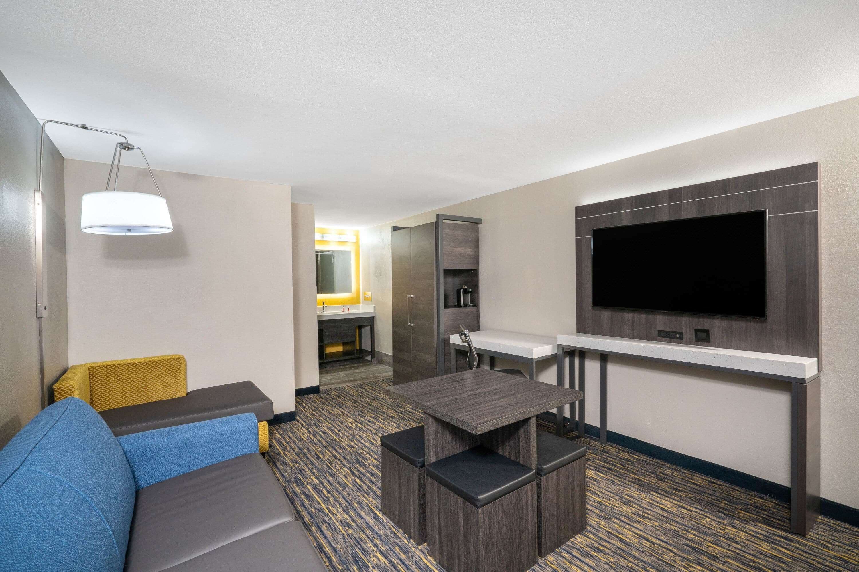 A hotel room with a sofa, a small table, a TV mounted on the wall, and a kitchenette in the background.