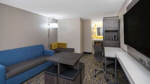 A hotel room with a sofa, a small table, a TV, and a kitchenette area.