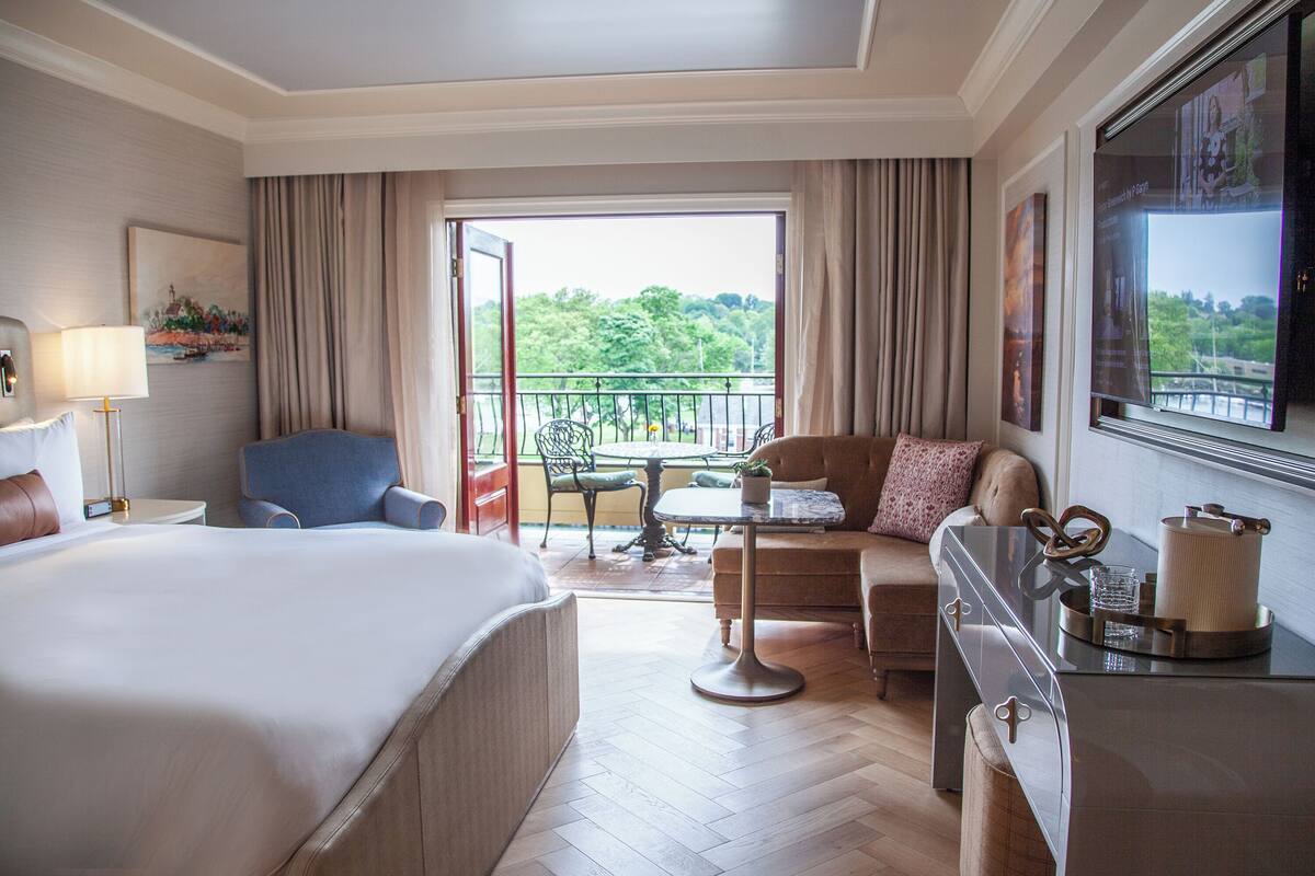 A hotel room with a bed, a sofa, a desk, a chair, a balcony with a table and chairs, and a TV.