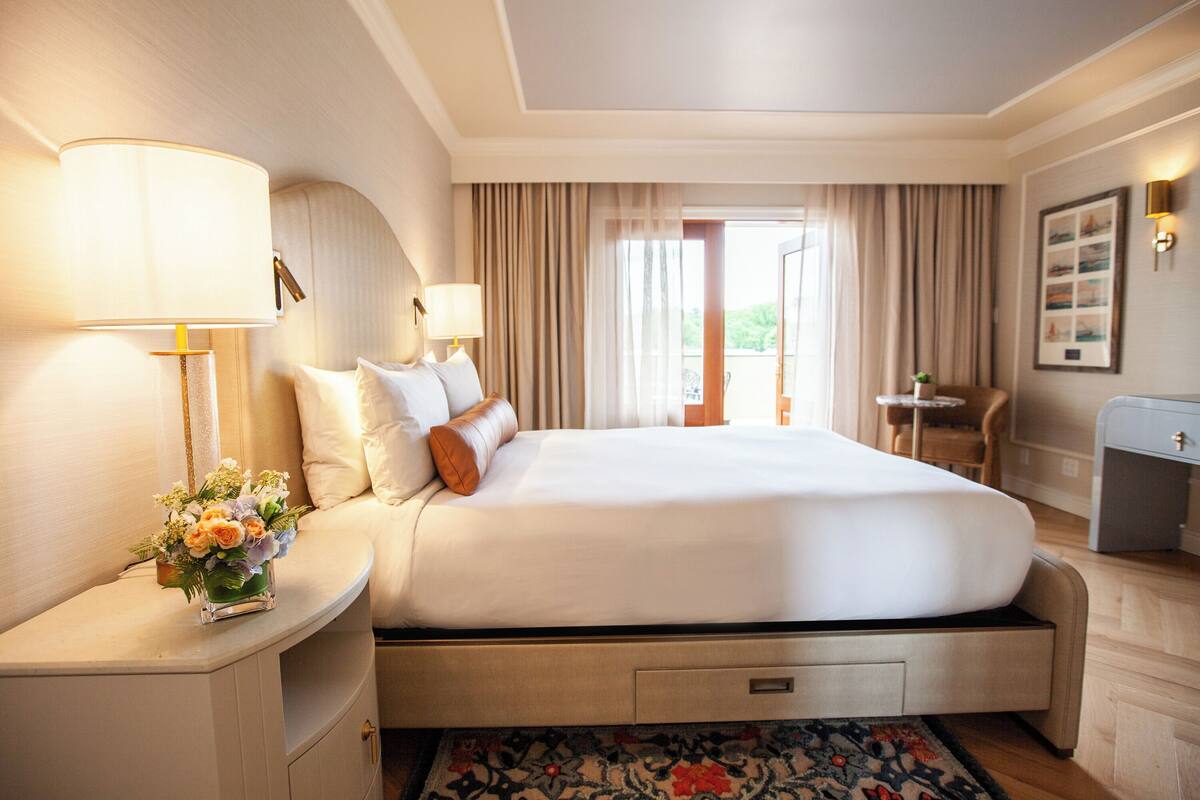 A hotel room with a bed, bedside lamps, a small table, a chair, a desk, and a window with curtains.
