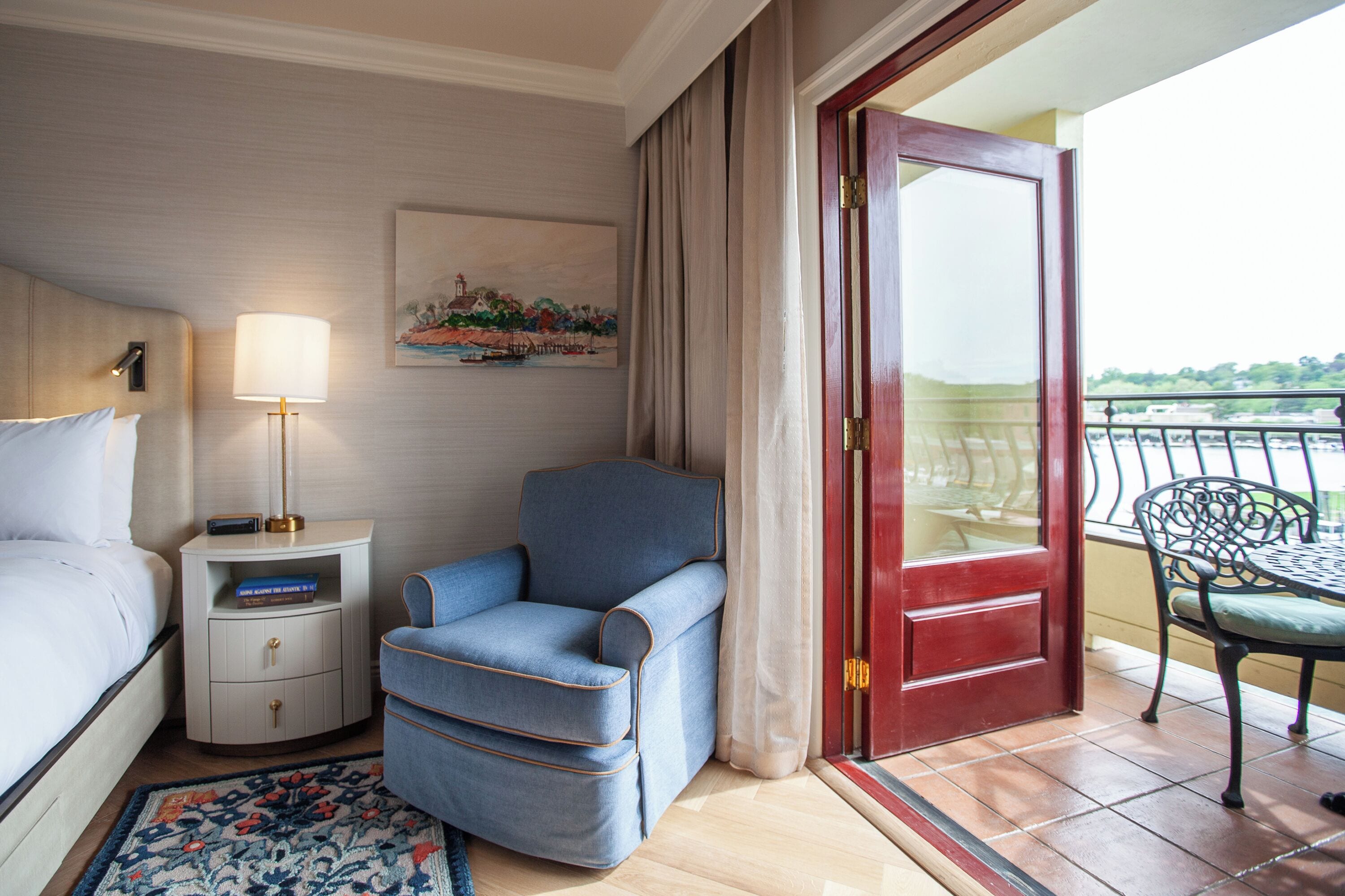 A hotel room with a bed, a nightstand with a lamp, a blue armchair, a red door, and a balcony with a chair.