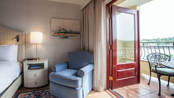 A hotel room with a bed, a nightstand with a lamp, a blue armchair, a red door, and a balcony with a chair.