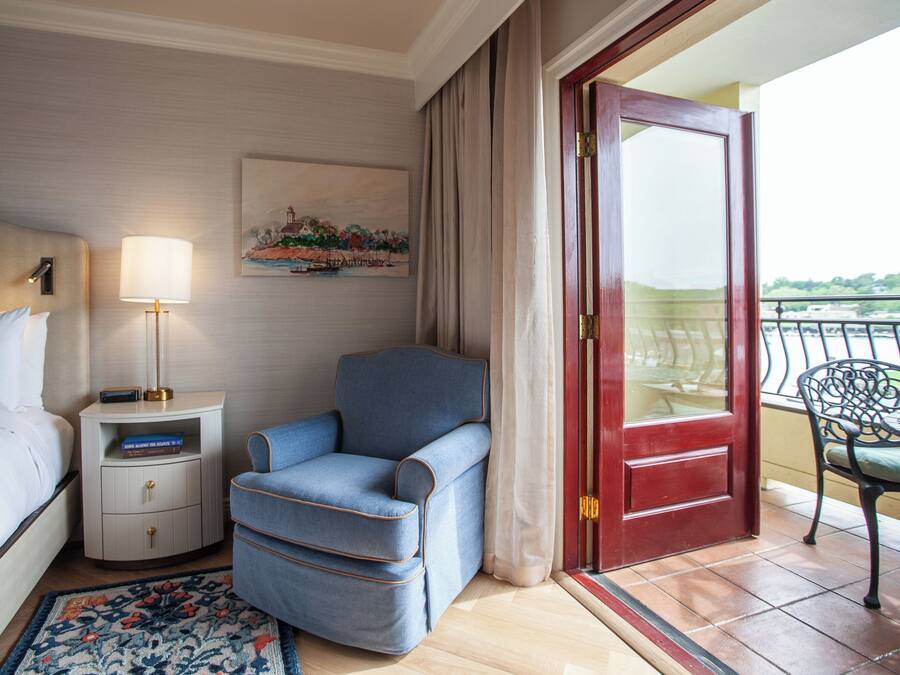 A hotel room with a bed, a nightstand with a lamp, a blue armchair, a red door, and a balcony with a chair.