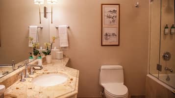Een badkamer met een marmeren wastafel, twee wandlampen, een lijst met een foto, een toilet en een douche met glazen wanden.