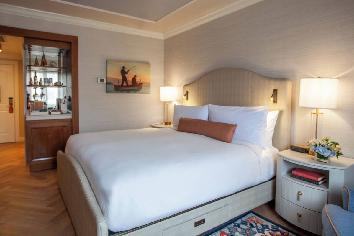 A hotel room with a large bed, bedside lamps, a nightstand with a radio and books, a small table with a vase, and a painting on the wall.