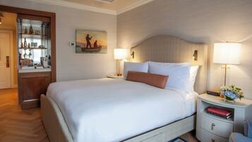 Habitación de hotel con una cama grande, lámparas de noche, una mesita de noche con una radio y libros, una pequeña mesa con un florero y un cuadro en la pared.