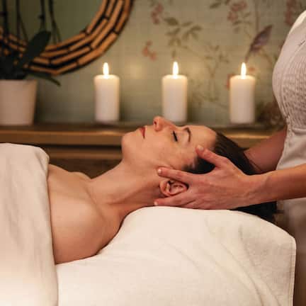 A person receiving a facial treatment in a room with a mirror, candles, and a potted plant.