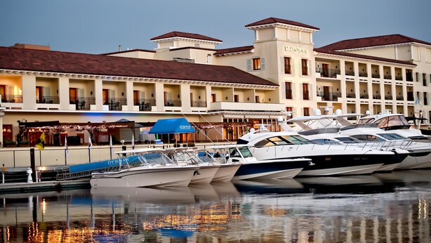 Un port de plaisance où sont amarrés plusieurs yachts, devant un hôtel doté de nombreux balcons et d’une tour centrale.