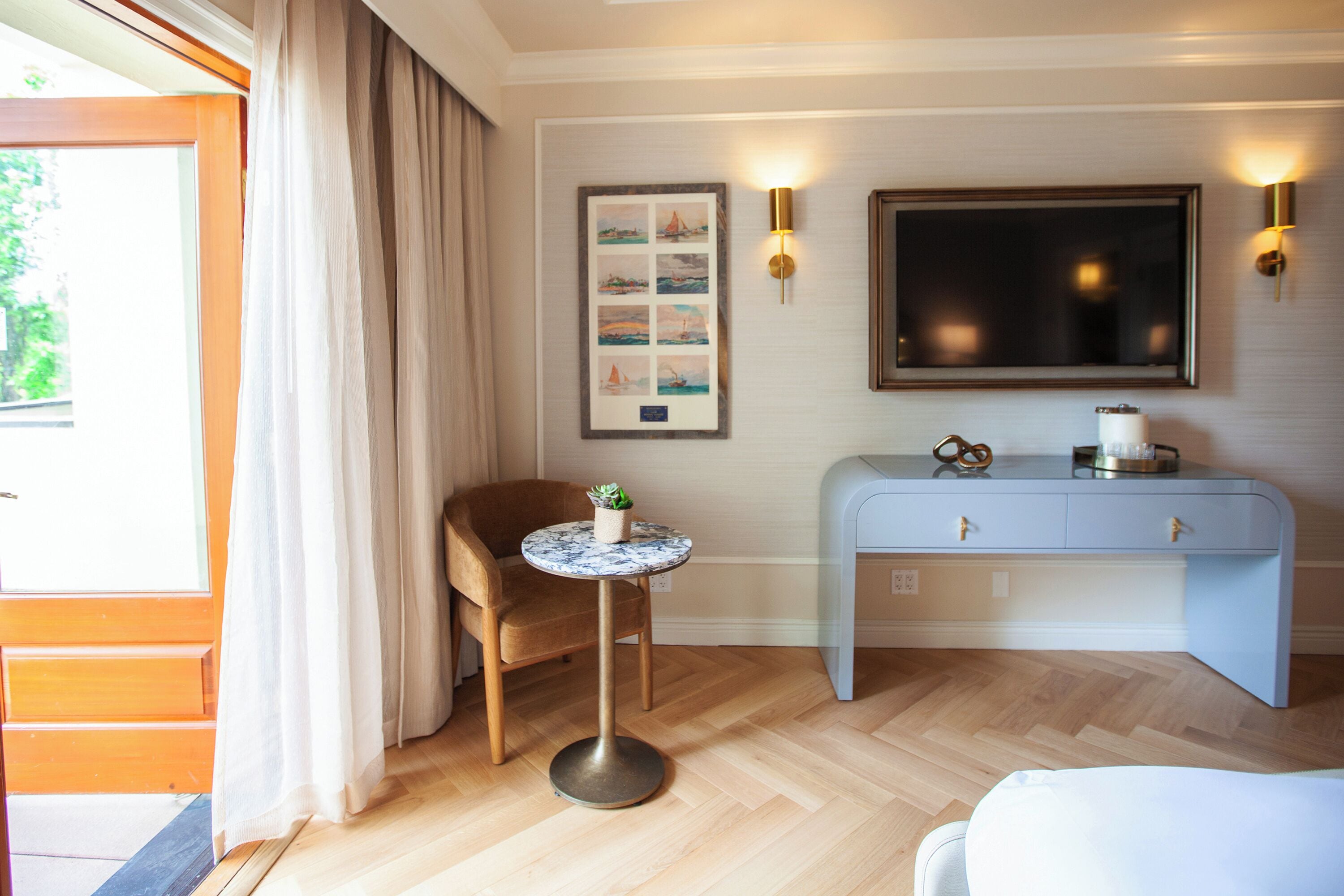 Une chambre d’hôtel avec un plancher en bois, une table ronde ornée d’une plante, un bureau bleu et un tableau encadré accroché au mur.