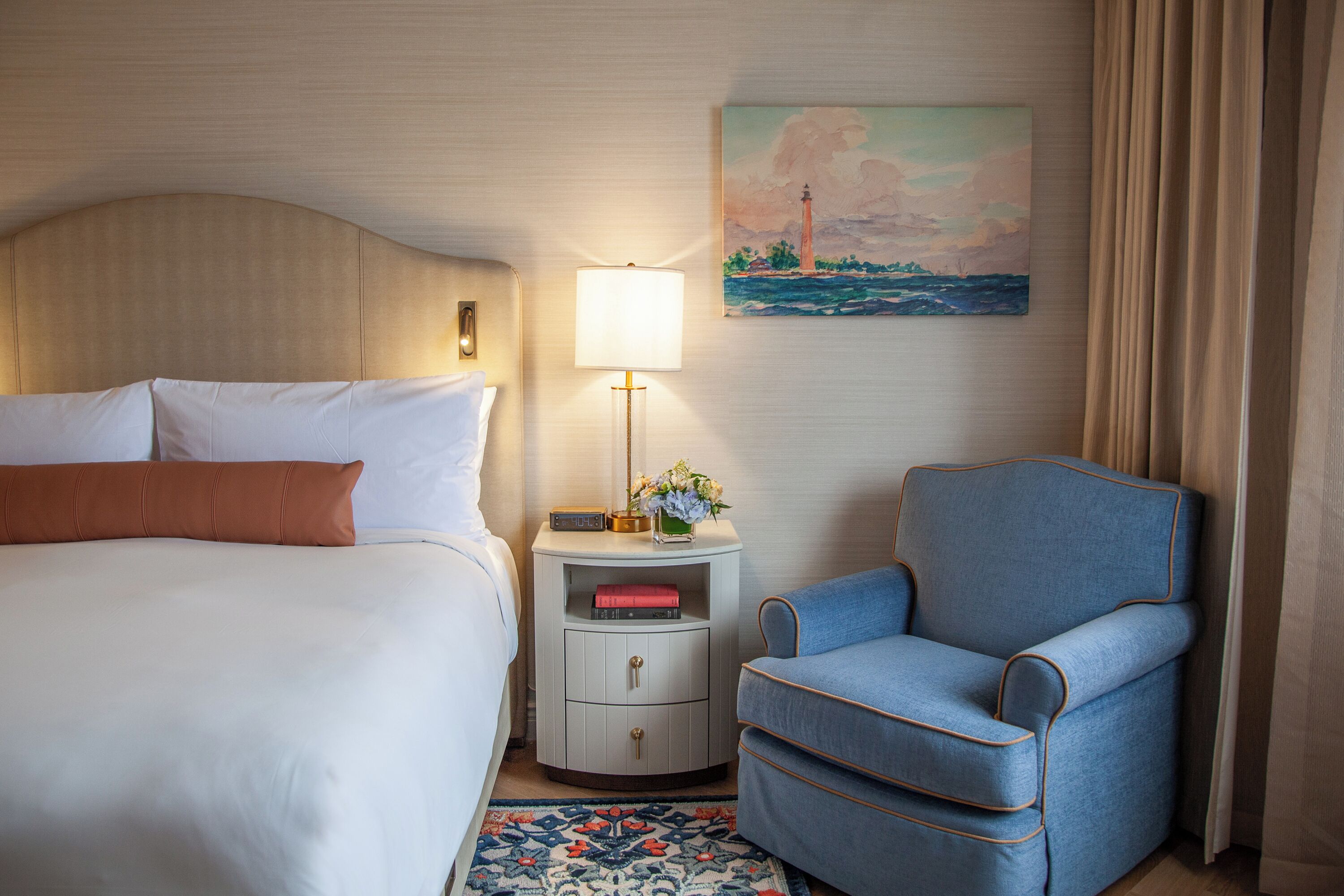 A hotel room with a bed, a nightstand with a lamp, a chair, and a painting on the wall.