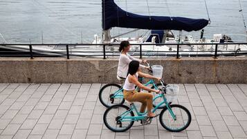 Duas pessoas andando de bicicleta azul-turquesa em uma orla pavimentada, com um barco atracado ao fundo.