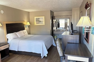 Una habitación de hotel con cama, mesita de noche, lámpara, silla, escritorio y una pequeña cocina.
