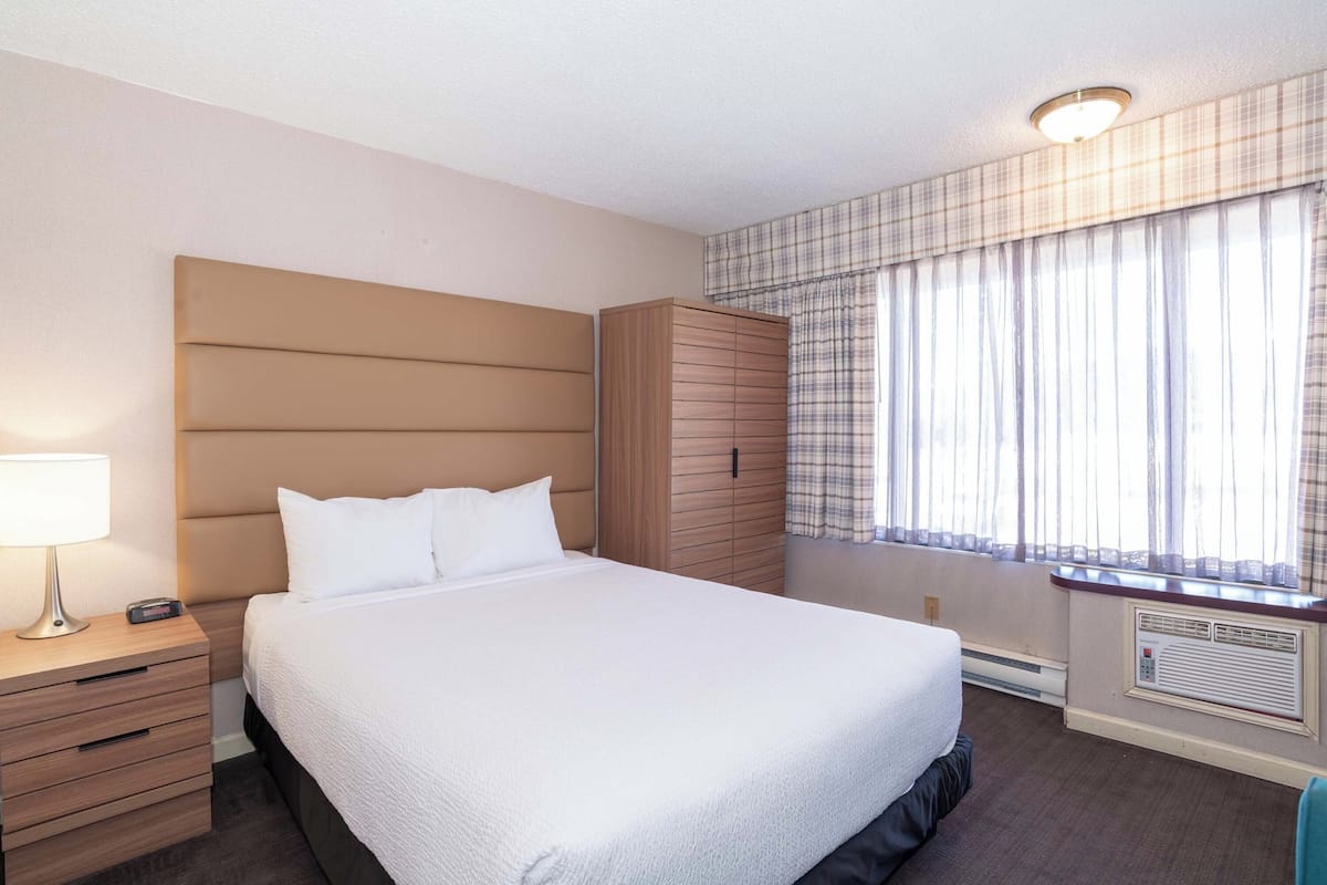 A neatly arranged hotel room with a large bed, a bedside table with a lamp, a wardrobe, and a window with curtains.