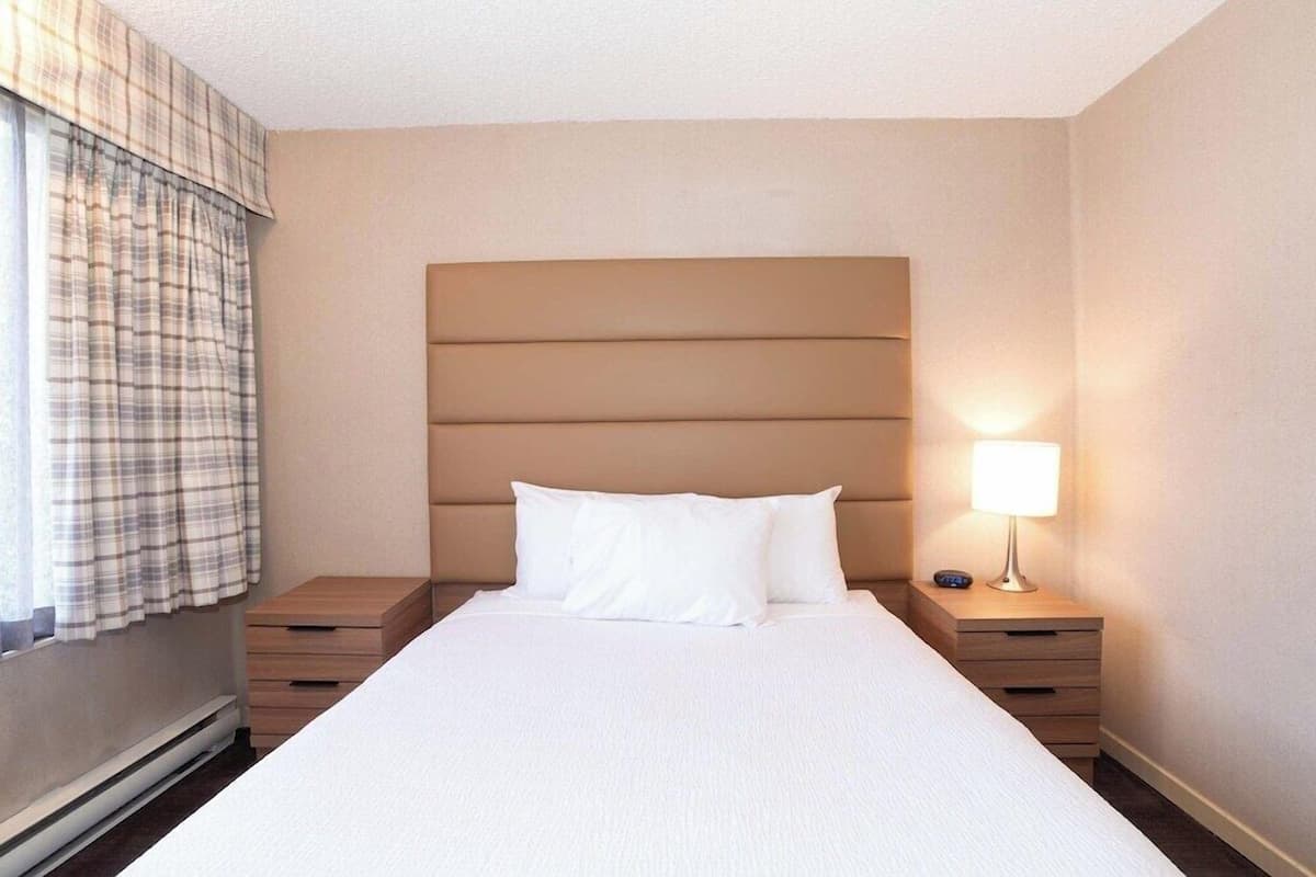 A hotel room with a large bed, a headboard, a bedside table with a lamp, and a window with curtains.