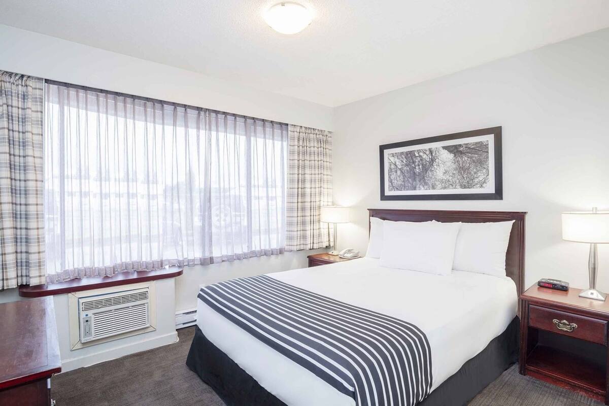 A hotel room with a large bed, two bedside tables with lamps, a window with sheer curtains, a radiator, and a framed picture on the wall.