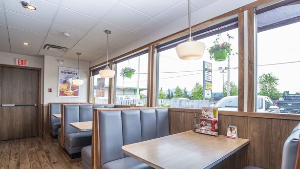 A restaurant interior with booths and tables, wooden flooring, hanging plants, and a view of the street outside.