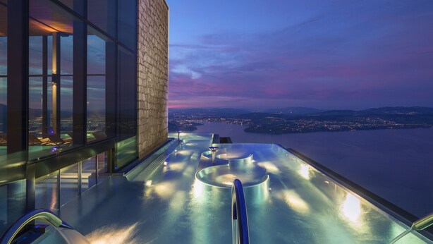 A rooftop infinity pool with a glass balustrade, a stone wall, and a cityscape view at dusk.