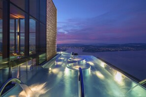 A rooftop infinity pool with a glass balustrade, a stone wall, and a cityscape view at dusk.