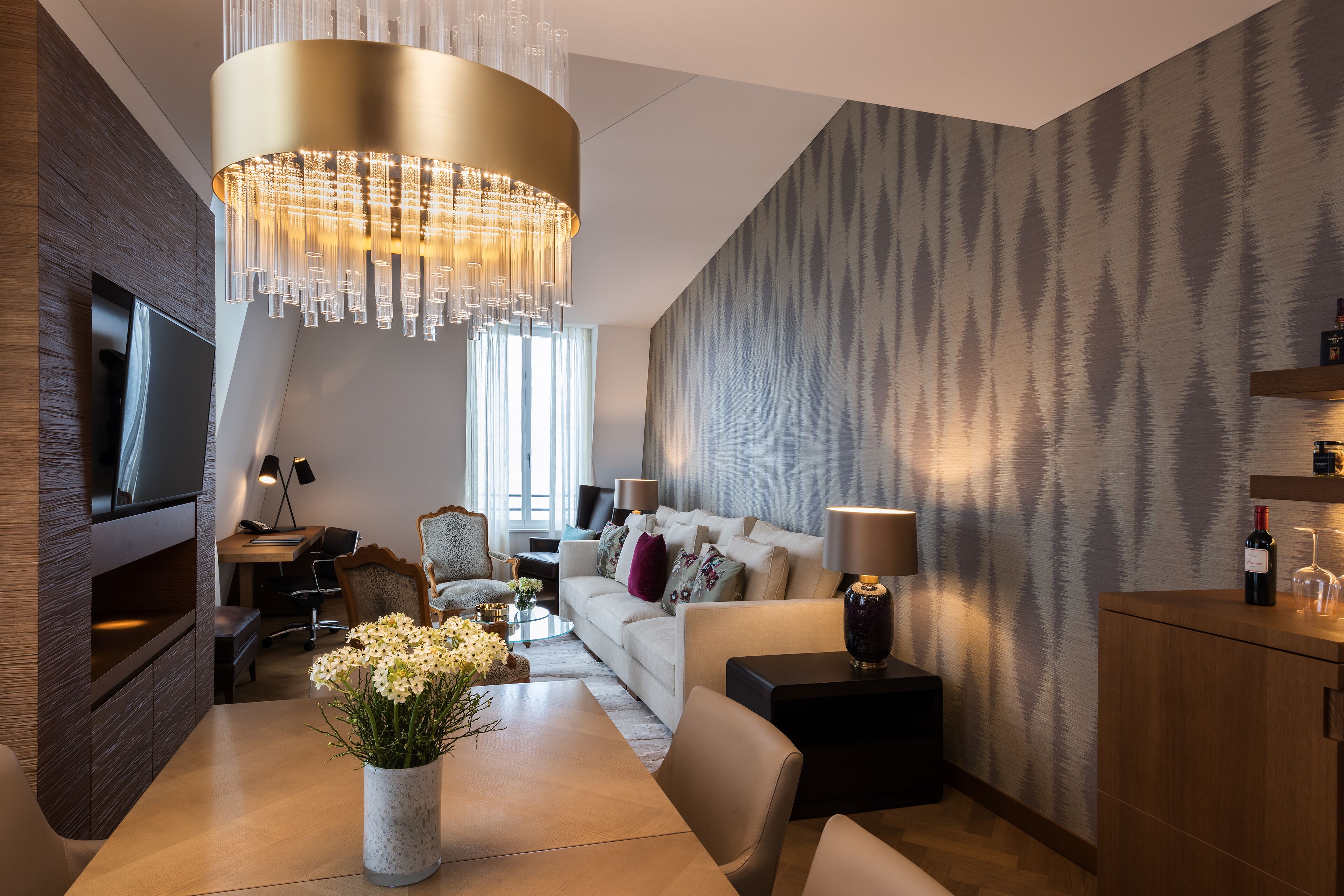 Un salon moderne avec un grand lustre, un ensemble de canapés, une table basse agrémentée de fleurs et un téléviseur fixé au mur.
