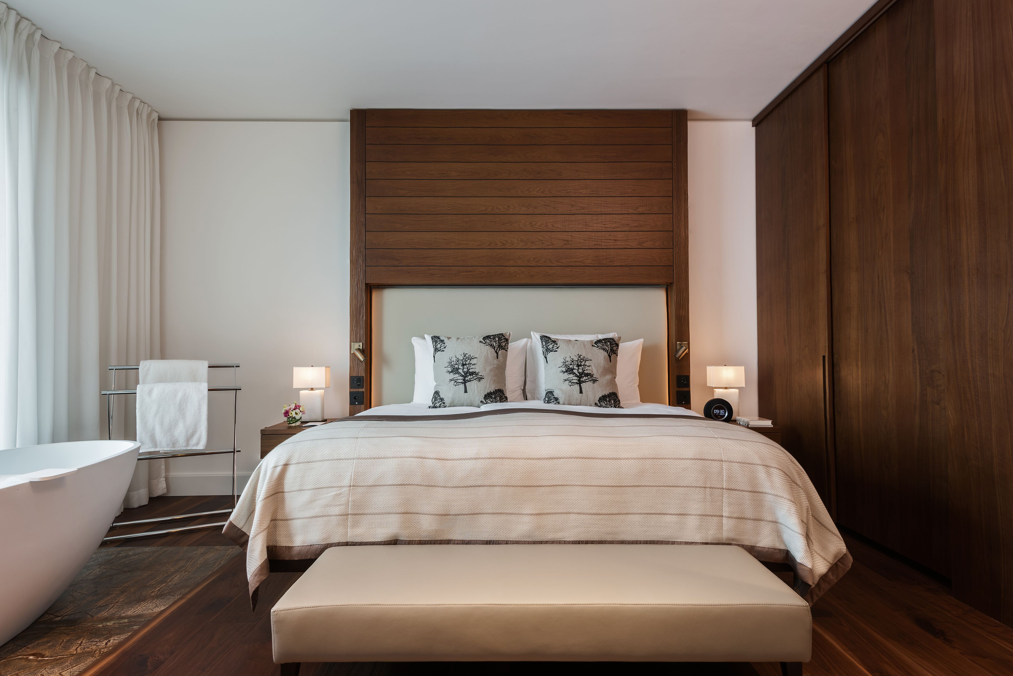 Ein modernes Hotelzimmer mit einem großen Bett, Nachttischlampen, einem Holzkopfschild und einem Badezimmer mit Badewanne und Handtuchhalter.