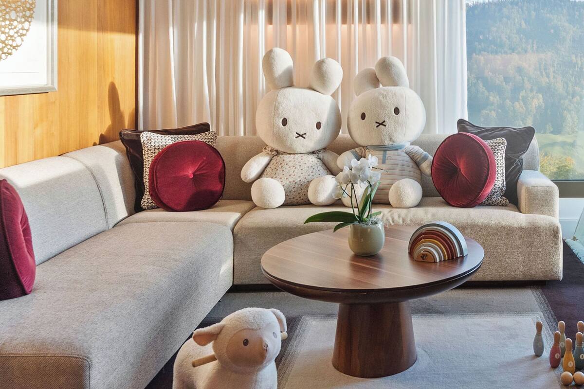 Uma sala de estar moderna com um sofá, dois coelhos de pelúcia brancos, um cavalinho de balanço e uma mesa de centro com um vaso e objetos decorativos.