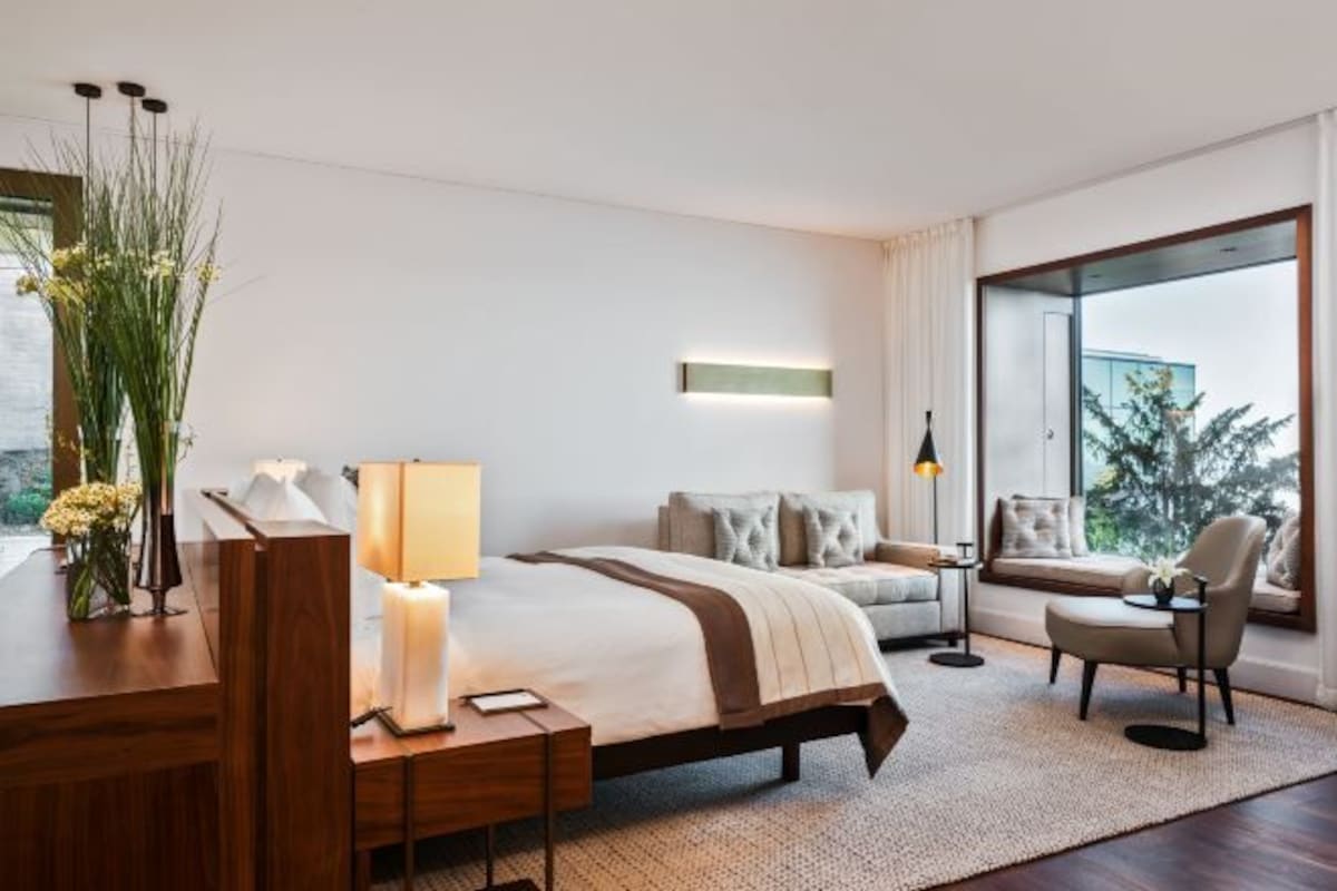 A modern hotel room with a large bed, a desk with a lamp, a chair, and a view of the outdoors through a large window.