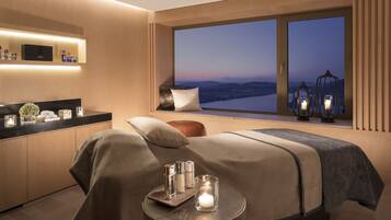 Habitación de hotel moderna con un gran ventanal que ofrece vistas de la ciudad al anochecer, una cama bien hecha, una mesita con velas y un minibar.