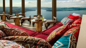 A luxurious room with a large window overlooking a lake and mountains, furnished with patterned cushions and traditional decor.