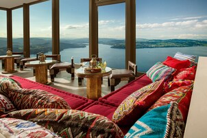 Una habitación lujosa con un amplio ventanal que da a un lago y montañas, decorada con cojines estampados y una decoración tradicional.