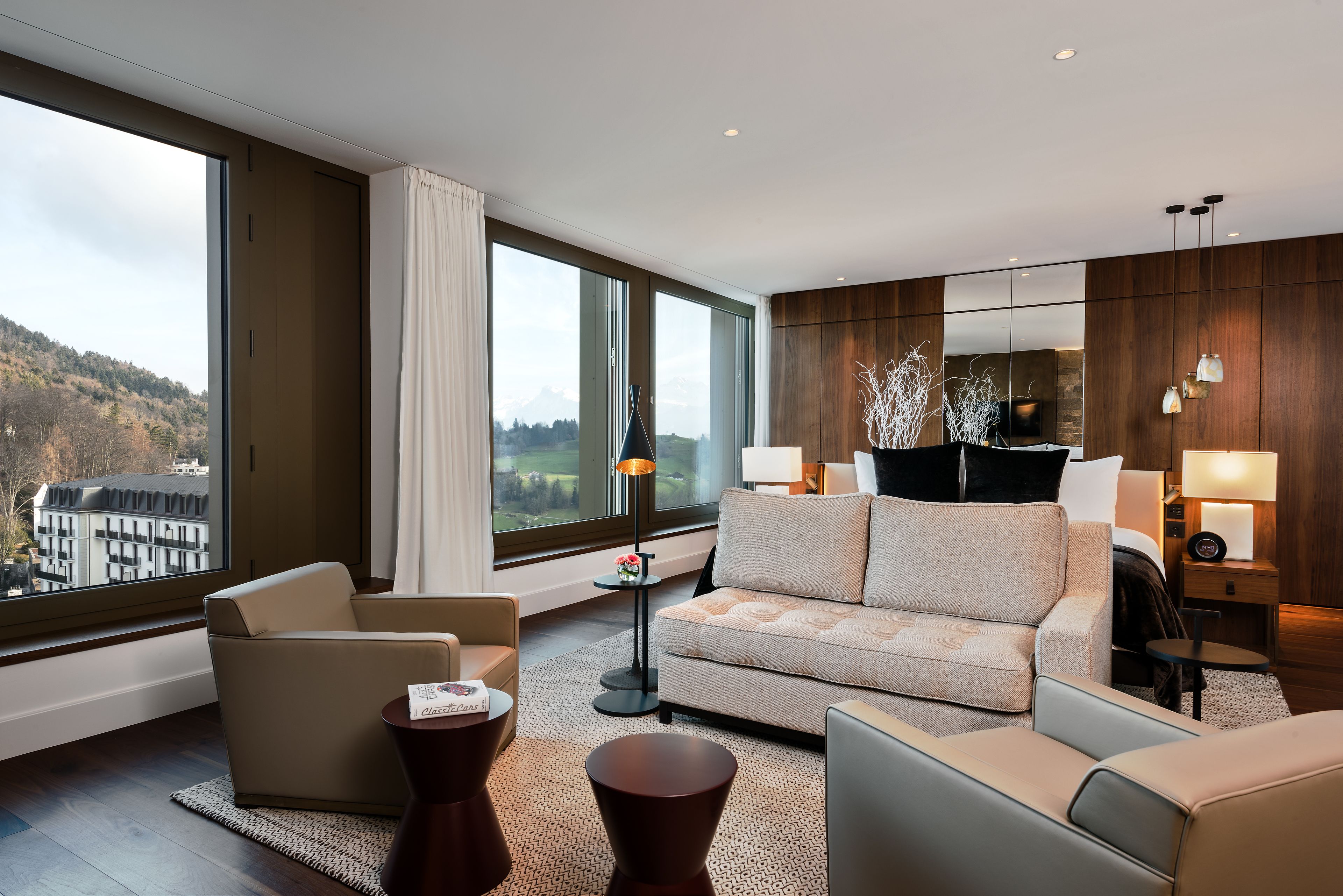 Ett modernt hotellrum med ett stort fönster som erbjuder utsikt över en stad och berg. Rummet innehåller en soffa, en fåtölj och ett litet bord med en lampa.