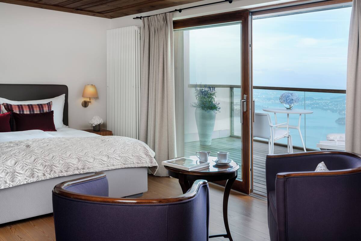 Quarto de hotel com uma cama, uma mesinha com uma chávena e um livro, duas cadeiras e uma varanda com vista para o mar.