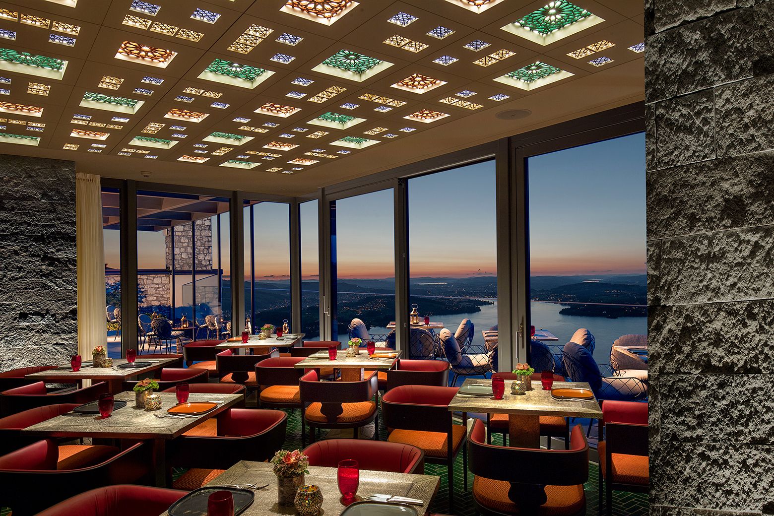 A restaurant with a high ceiling featuring stained glass windows, large windows offering a view of a river and distant landscape, and red upholstered chairs.