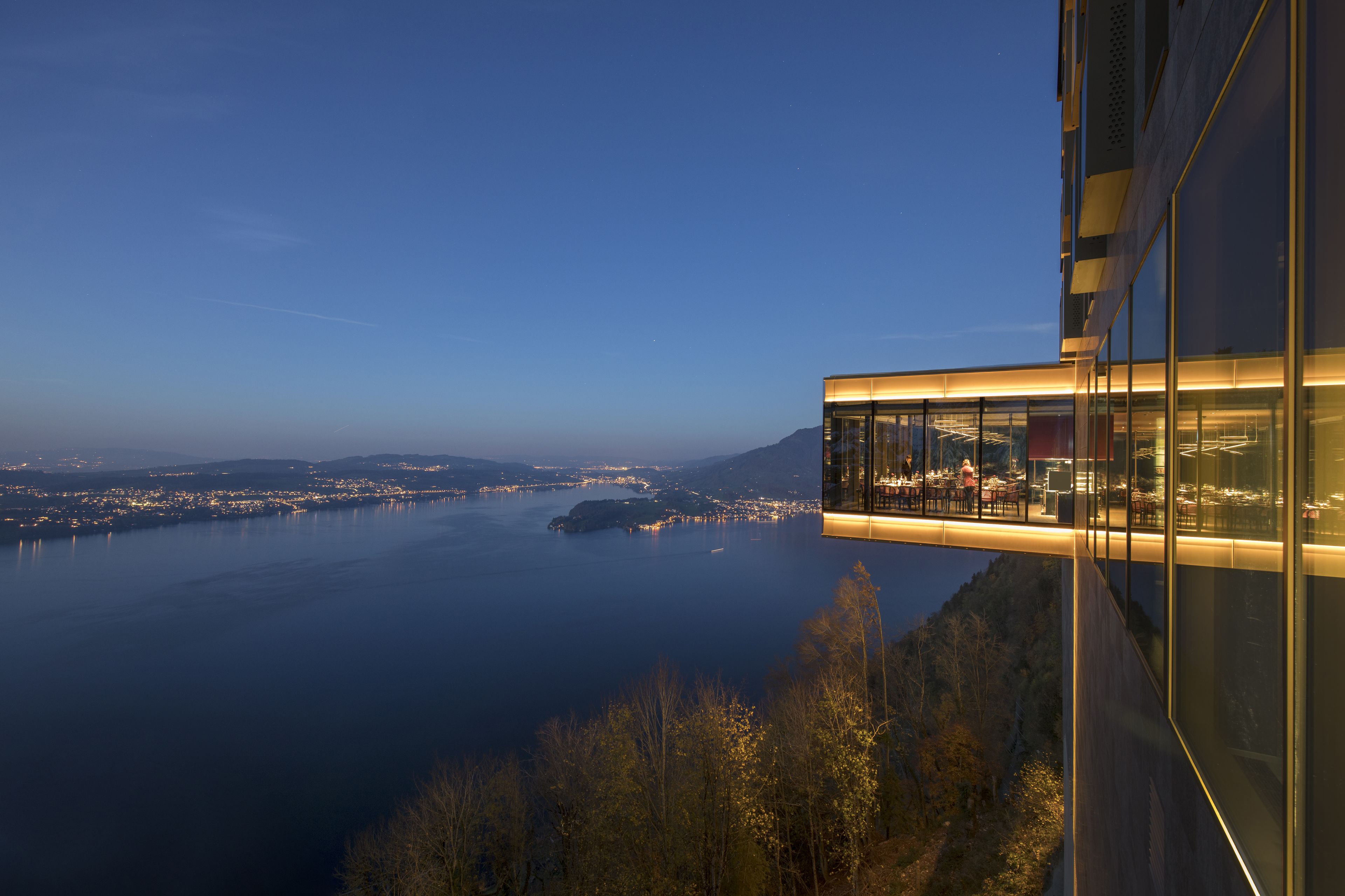 A modern building with a glass facade overlooking a body of water and a distant cityscape.