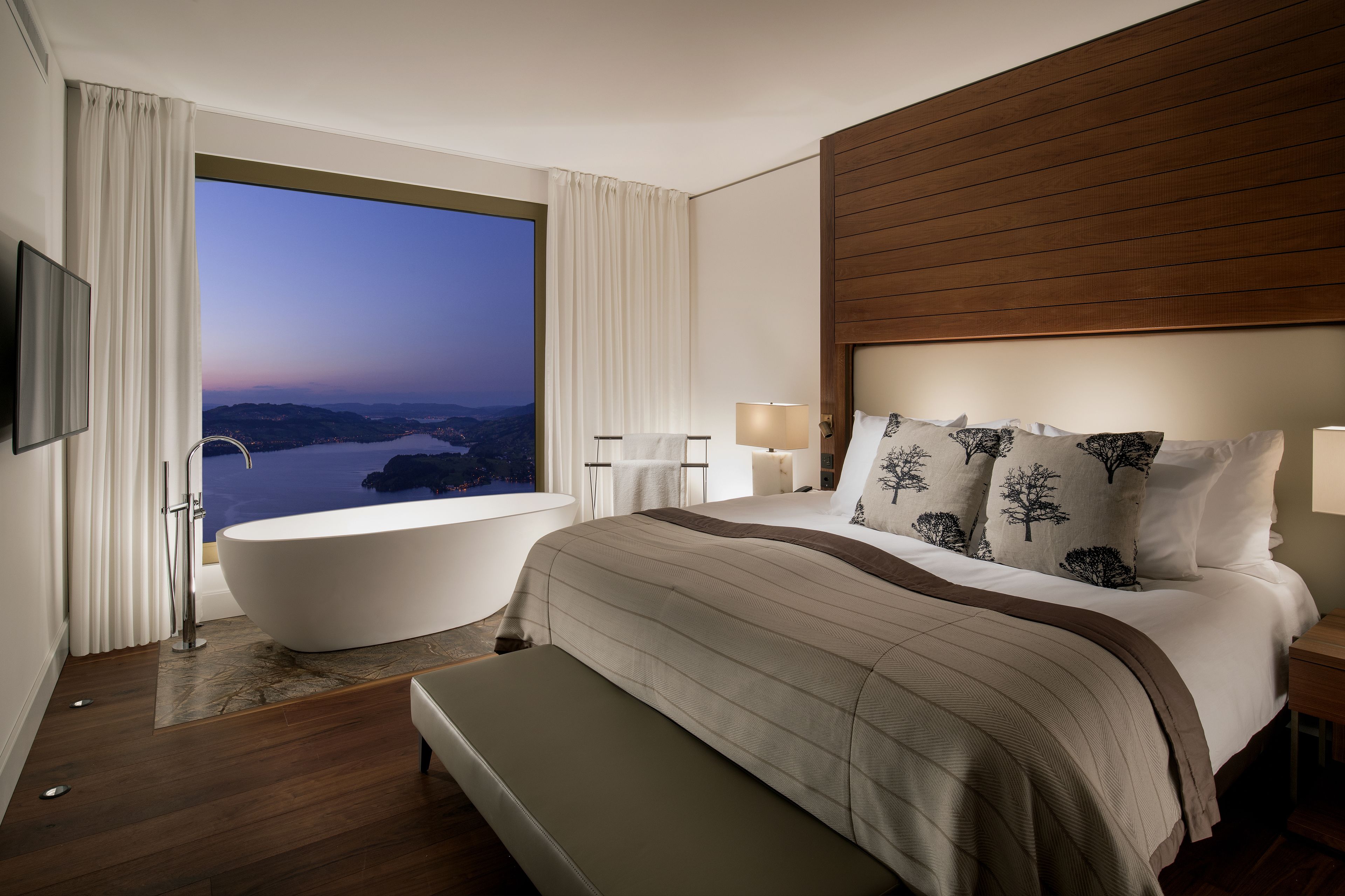 A modern bedroom with a large window, a bathtub, a bed with a headboard, and a bench.