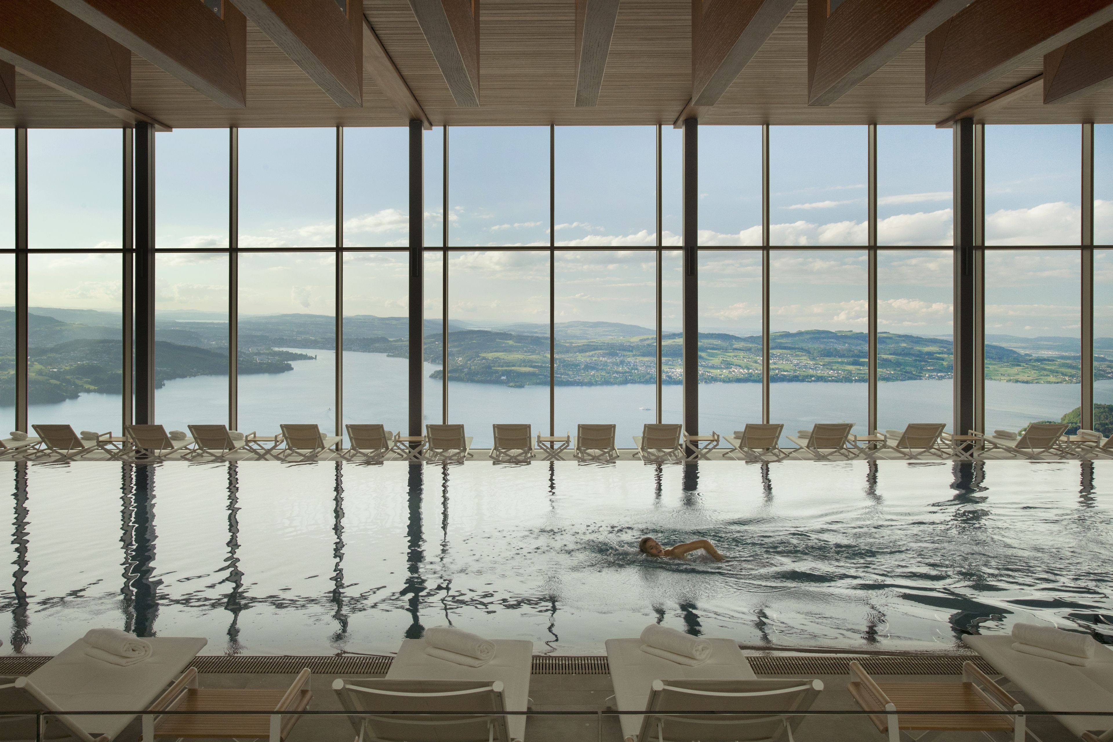 An indoor swimming pool with clear views of a lake and mountains through large windows.
