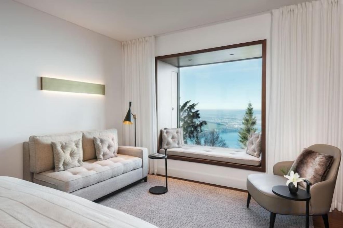 A modern hotel room with a large window, a sofa, a chair, and a bed.