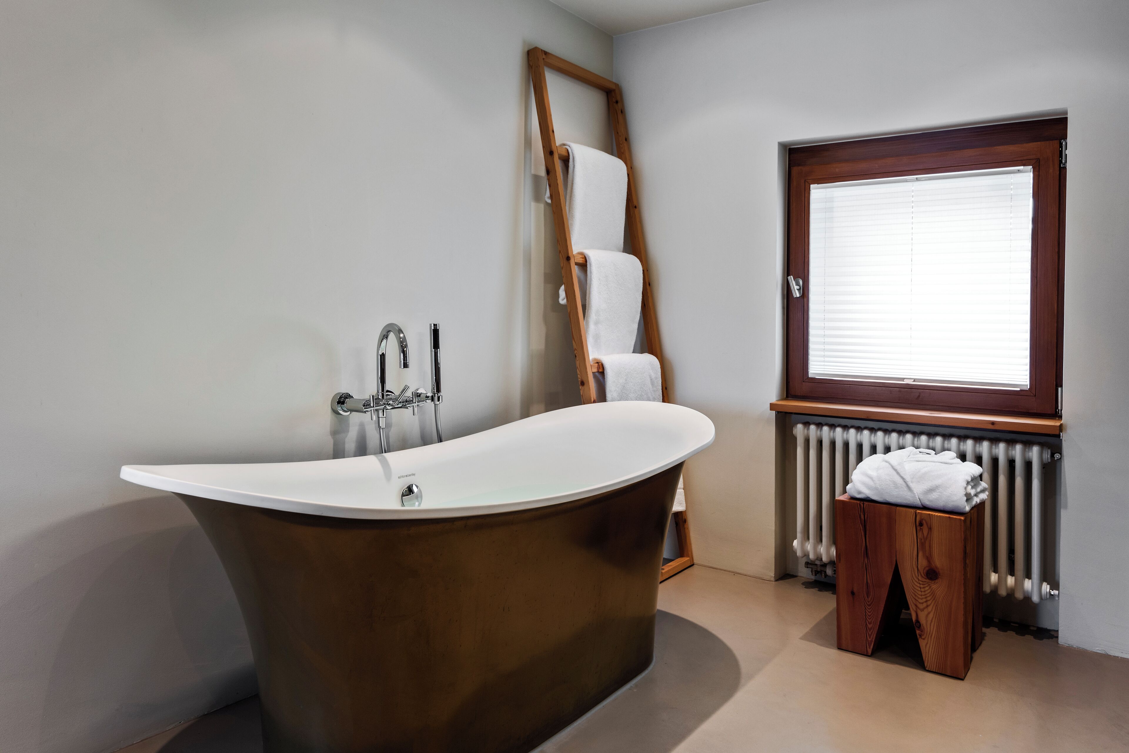 Ein modernes Badezimmer mit einer freistehenden Badewanne, einer Holzleiter, einem Fenster mit Jalousien und einem Heizkörper.