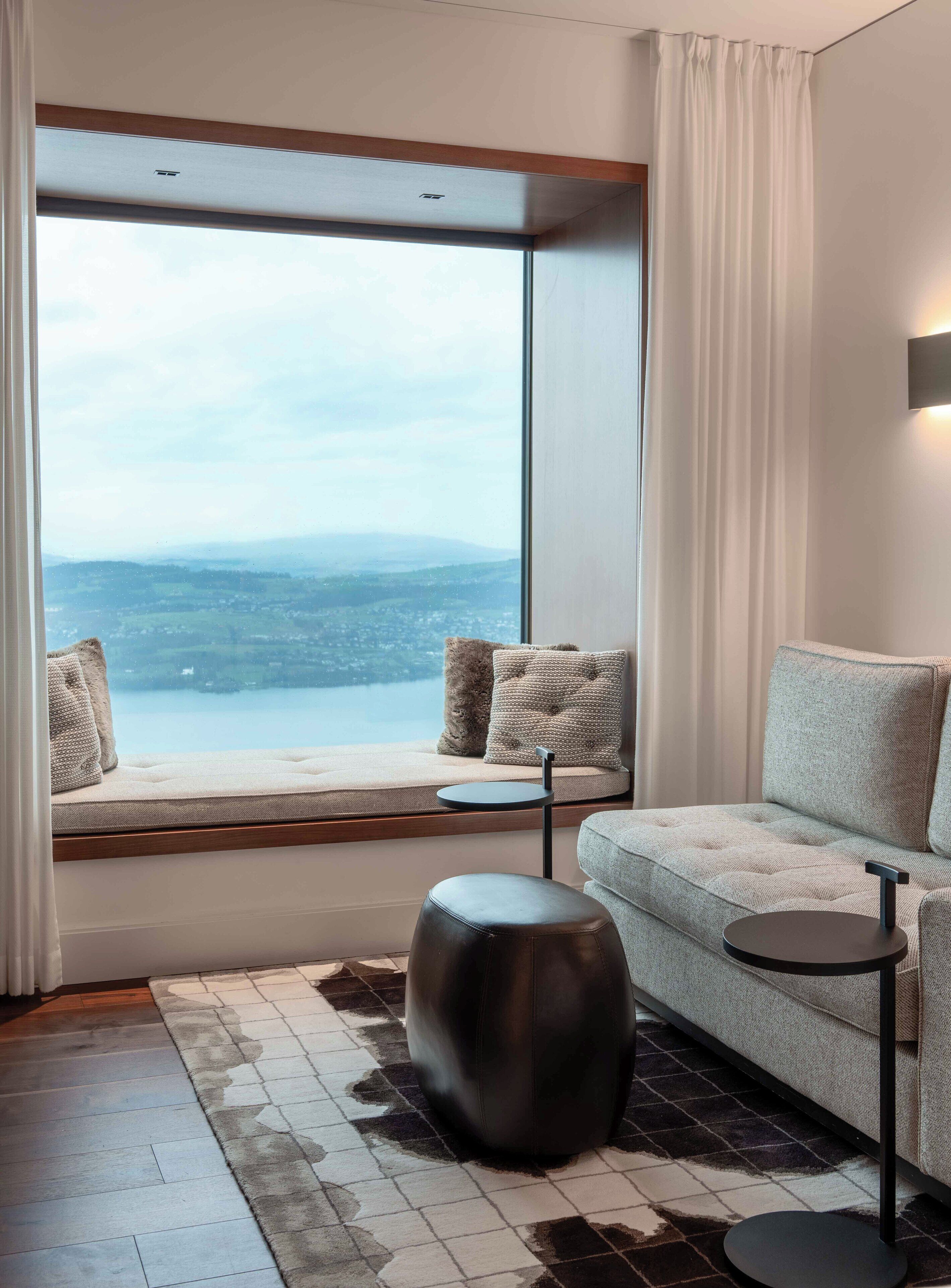A modern living room with a large window, a sofa, a round ottoman, and a small side table.