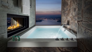 A bathroom with a bathtub, a fireplace, and a view of a cityscape at dusk.