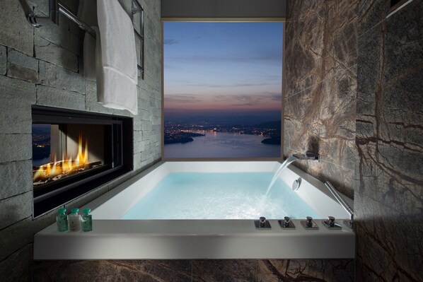 A bathroom with a bathtub, a fireplace, and a view of a cityscape at dusk.