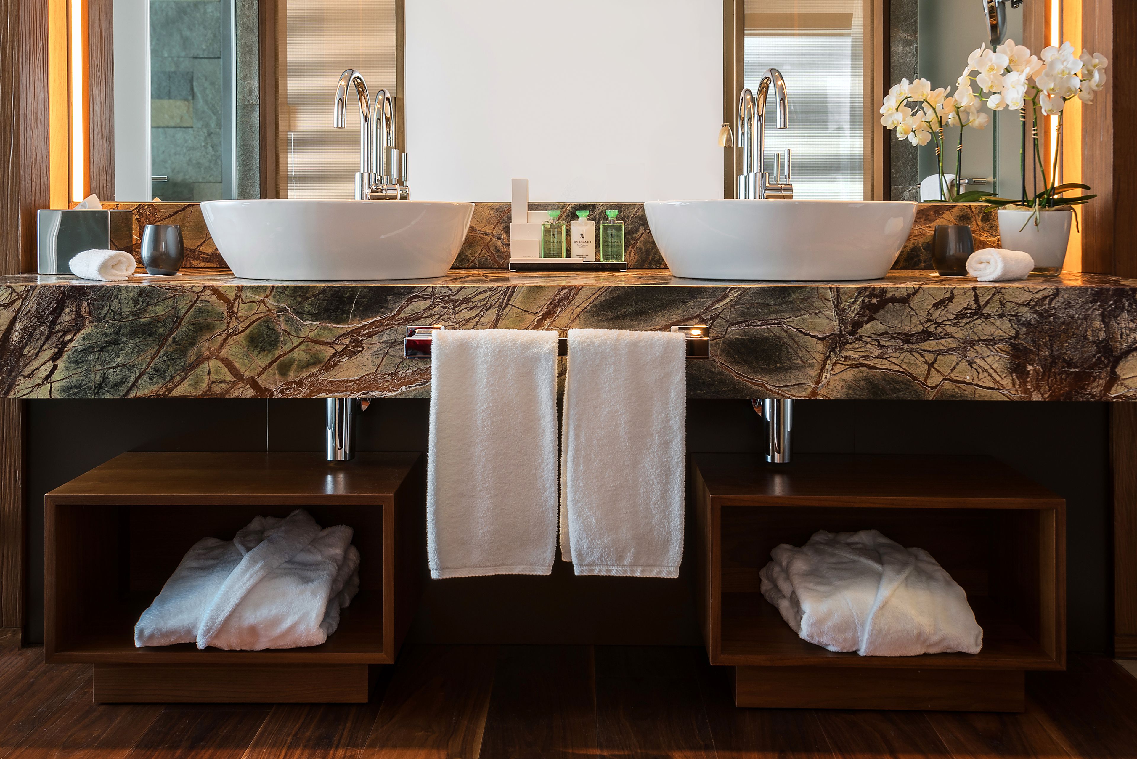 Ein Badezimmer mit zwei großen weißen Waschbecken, einer Marmorarbeitsplatte und einem Spiegel darüber. Es gibt zwei Handtuchhaltern mit aufgehängten Handtüchern, sowie ein Regal mit Flaschen und einer Blumenvase.