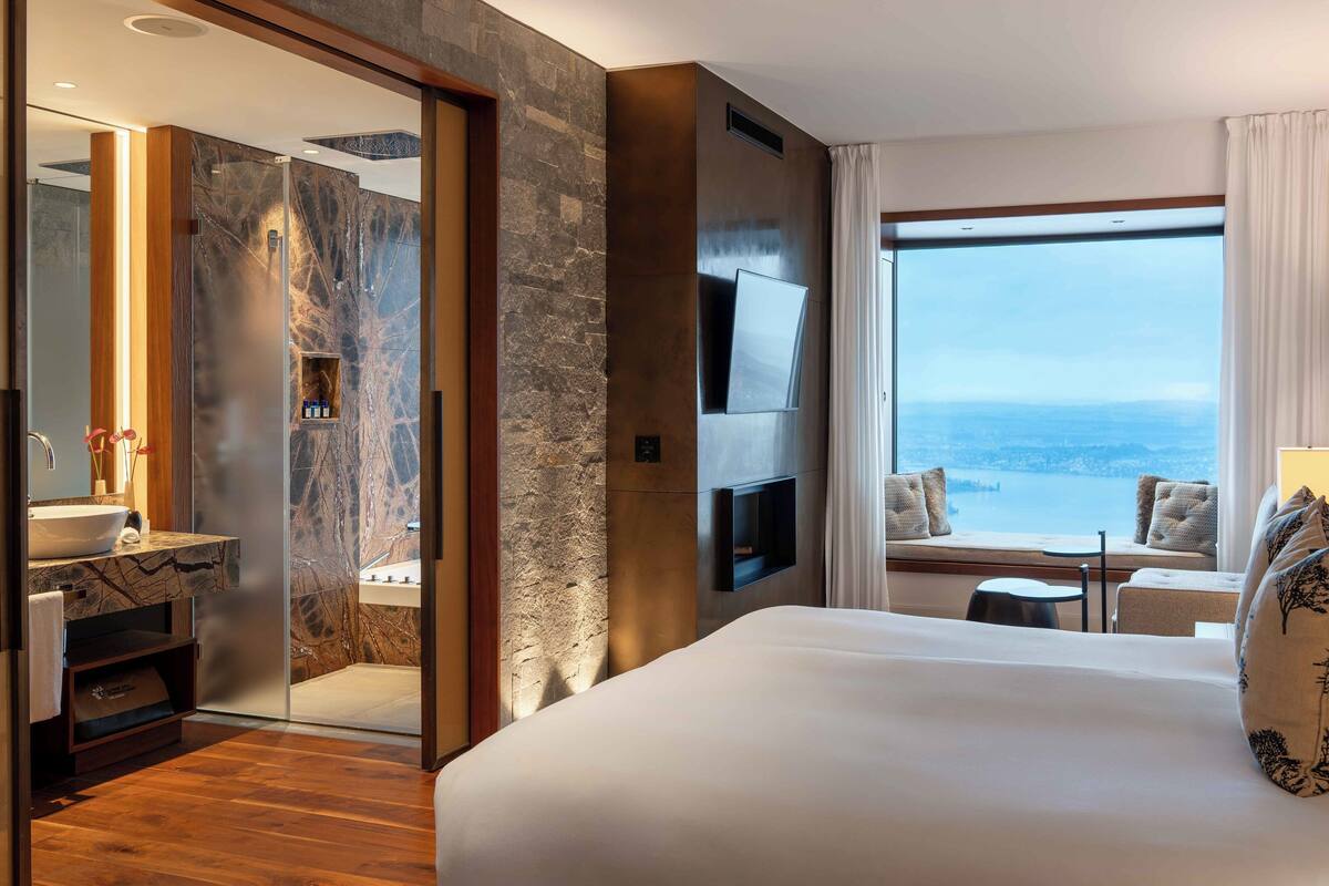 Quarto de hotel moderno com uma cama grande, banheiro com toucador de mármore, televisão fixada na parede e vista para a cidade através de amplas janelas.